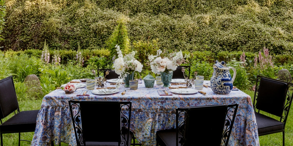 a table set with flowers vases pitches plates and glasses outside in a garden with six black chairs surrounding